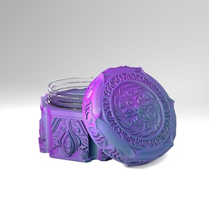 Può includere: Un barattolo iridescente viola e blu con coperchio abbinato, caratterizzato da intricati motivi in rilievo di foglie e forme geometriche. Il contenitore presenta un bordo interno in vetro trasparente e una superficie esterna testurizzata.