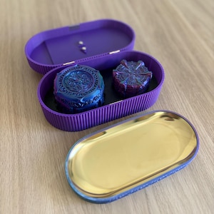 May include: An oval purple storage box with a ribbed exterior and a hinged lid. Inside, two circular blue and purple patterned containers sit on a black base. A separate oval tray with a reflective gold interior and a blue exterior is placed in front.