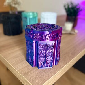 Pode incluir: Um recipiente hexagonal com um acabamento iridescente que alterna entre o roxo profundo e o azul vibrante. O exterior apresenta padrões botânicos intrincados em relevo em cada lado e um design geométrico detalhado na tampa.