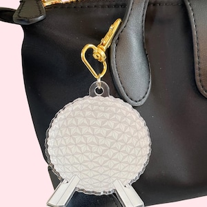 May include: A clear acrylic keyring with a geometric design, attached to a black handbag with a gold-coloured clasp. The keyring has a detailed, spherical shape with a pattern of interconnected triangles.