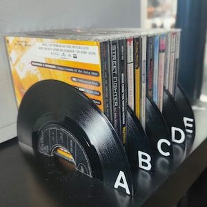 May include: A black vinyl record bookend with the letters A, B, C, D, and E on the side. The bookend is holding up a stack of CDs with titles like "Street Fighter", "Music from the Motion Picture Pulp Fiction", and "Various Artists - New Jersey".