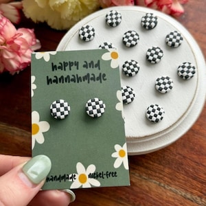 May include: A pair of black and white checkered stud earrings on a green card with the text "happy and hannahmade" and "handmade nickel-free". Additional checkered earrings are displayed on a white surface, surrounded by flowers.