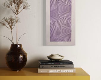 May include: A rectangular, lavender-colored wall art piece with a repeating semi-circular pattern. A brown vase with dried flowers sits on a yellow table, along with a stack of books and a small white bowl.