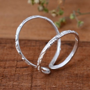 以下が含まれることがあります： 複雑な花の模様が施された銀の指輪2つ。指輪は絡み合っており、木製の表面の上に置かれています。