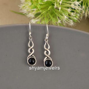 Può includere: Un paio di orecchini pendenti color argento con un design a spirale e una pietra nera rotonda alla base. Gli orecchini sono esposti su una superficie grigia, con il testo "shyamjewelrs" visibile.
