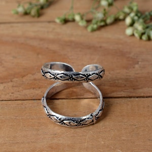 Könnte beinhalten: Ein silberfarbener Doppelring mit detailliertem geometrischem Muster. Der Ring wird auf einer Holzoberfläche präsentiert, die sein aufwendiges Design und seinen metallischen Glanz hervorhebt. Die Ringbänder sind verbunden und schaffen ein einzigartiges und stilvolles Accessoire.