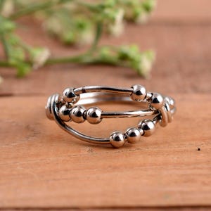 Könnte beinhalten: Ein silberfarbener Ring mit einem einzigartigen Design. Der Ring hat drei Bänder, wobei kleine, runde Perlen auf zwei davon aufgefädelt sind. Die Perlen sind gleichmäßig verteilt und verleihen dem Ring Textur. Der Ring ist vor einem Holzgrund platziert.