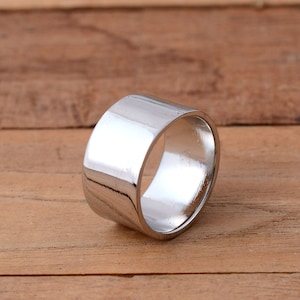 Peut inclure: Une large bague argentée polie avec un anneau plat. La bague est posée sur un fond en bois chaleureux, mettant en valeur sa surface réfléchissante. Le design est simple et moderne, adapté au quotidien. La largeur de la bague est d'environ 1 cm.