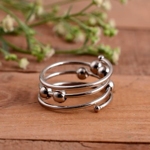 Könnte beinhalten: Ein silberfarbener Spiralring mit mehreren Bändern und kleinen, runden Kugeln. Der Ring wird auf einer Holzoberfläche mit einem verschwommenen Hintergrund aus Grünzeug präsentiert.