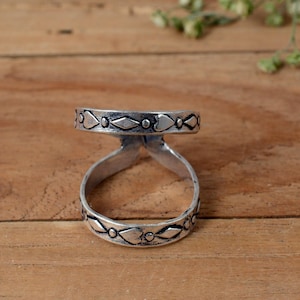 Puede incluir: Un anillo plateado con un diseño abierto. El anillo presenta una banda decorativa con recortes alternos en forma de diamante y corazón. El anillo está sobre una superficie de madera.