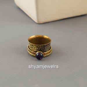 Pode incluir: Um anel dourado com uma faixa texturizada e uma faixa menor com um design trançado. Uma pequena gema redonda e roxa está engastada no anel. O anel está sobre uma superfície cinza. O texto "shyamjewelrs" está na parte inferior.