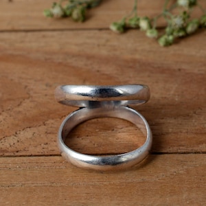 May include: A silver-colored ring with a unique double-band design. The ring features two connected bands, one above the other, creating a distinctive look. The metal has a slightly aged appearance, adding to its character. The ring is displayed on a wooden surface.