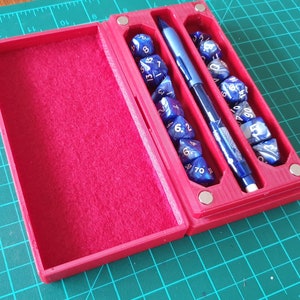 May include: A red dice box, open to reveal blue and white dice and a blue pen. The box has a felt lining and is on a green cutting mat. The box is approximately 15 cm long.