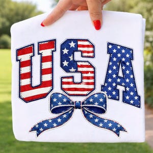Op de afbeelding: Wit kledingstuk met het woord "USA" in rood, wit en blauw. De letters zijn versierd met strepen en sterren, en een blauwe strik met witte sterren staat onderaan. Het kledingstuk wordt door een hand omhoog gehouden.