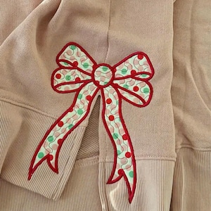 May include: Close-up of a pale pink sweatshirt with a large embroidered bow. The bow has a red outline with a pattern of red and green polka dots on a white background. The sweatshirt has ribbed cuffs.