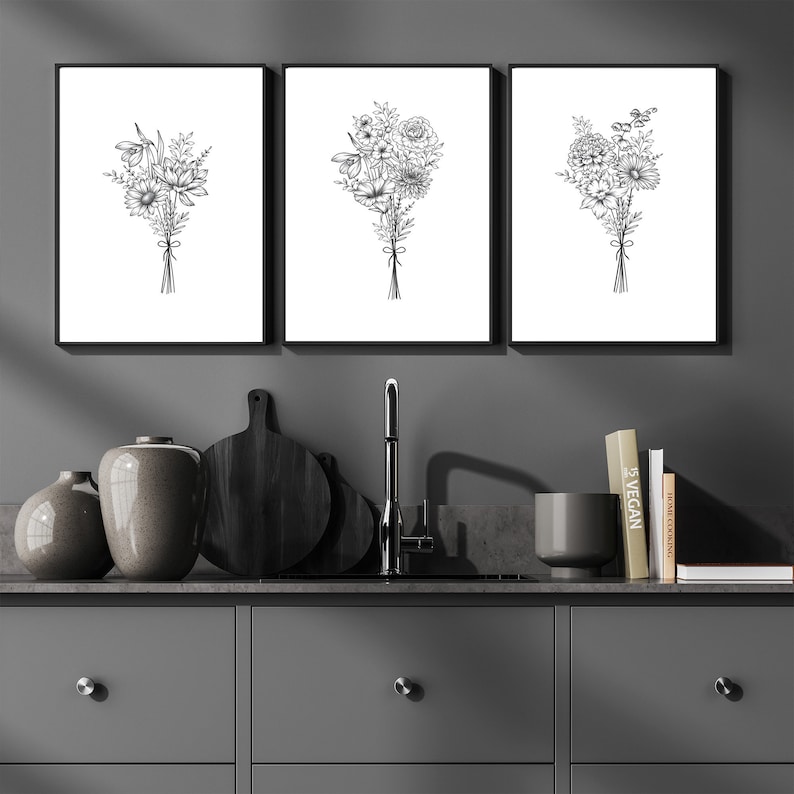 May include: Three black and white framed prints of floral bouquets, hanging on a gray wall above a kitchen counter with a sink, a cutting board, and vases.