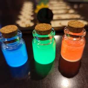 May include: Three small glass bottles with cork stoppers, each filled with a different colored liquid: blue, green, and orange. The liquids glow brightly, creating a vibrant and eye-catching display. The bottles are arranged on a dark surface.