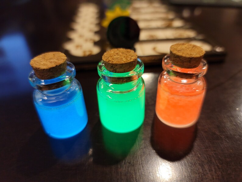 May include: Three small glass bottles with cork stoppers, each filled with a different colored liquid: blue, green, and orange. The liquids glow brightly, creating a vibrant and eye-catching display. The bottles are arranged on a dark surface.