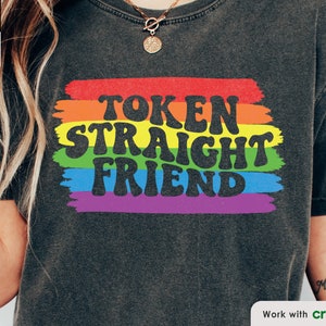 May include: A black t-shirt with the text "Token Straight Friend" in rainbow colors. The text is in a retro font style and is surrounded by a rainbow stripe design.