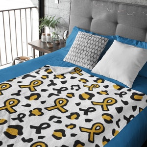 May include: A white fleece throw blanket with a black and gold leopard print pattern and yellow ribbon designs. The blanket is on a blue bed.