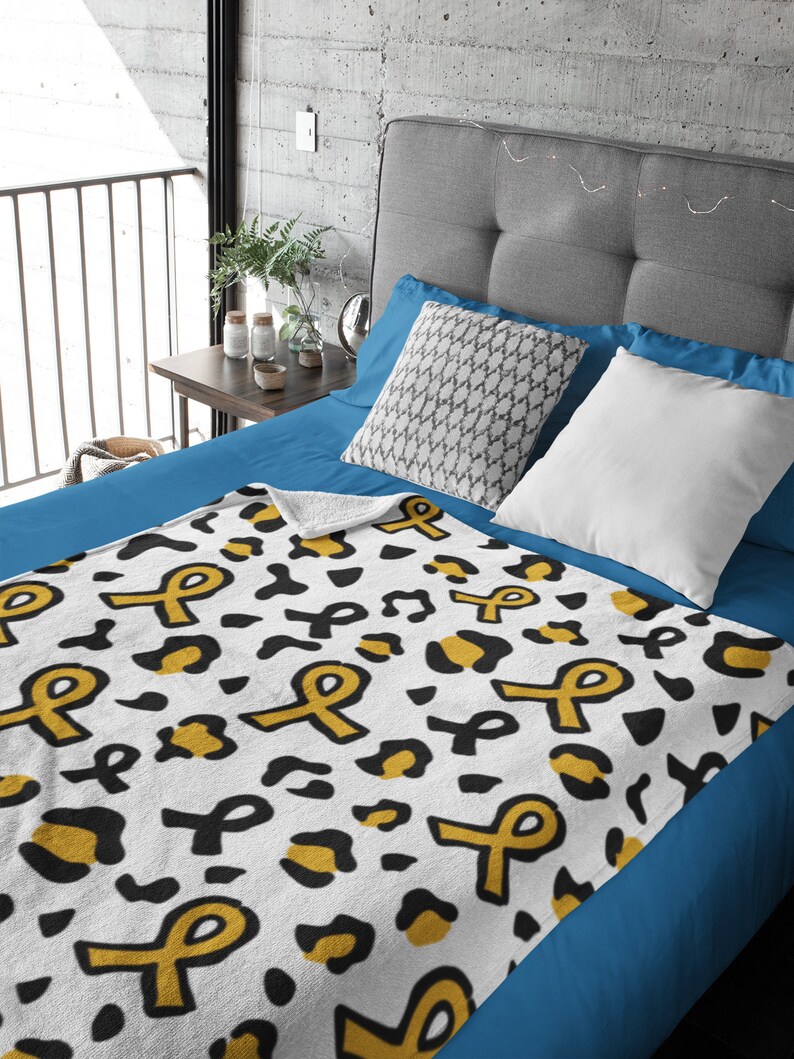 May include: A white fleece throw blanket with a black and gold leopard print pattern and yellow ribbon designs. The blanket is on a blue bed.