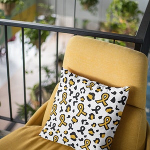 May include: A white throw pillow with a black and gold leopard print pattern. The pillow features a repeating design of yellow awareness ribbons.