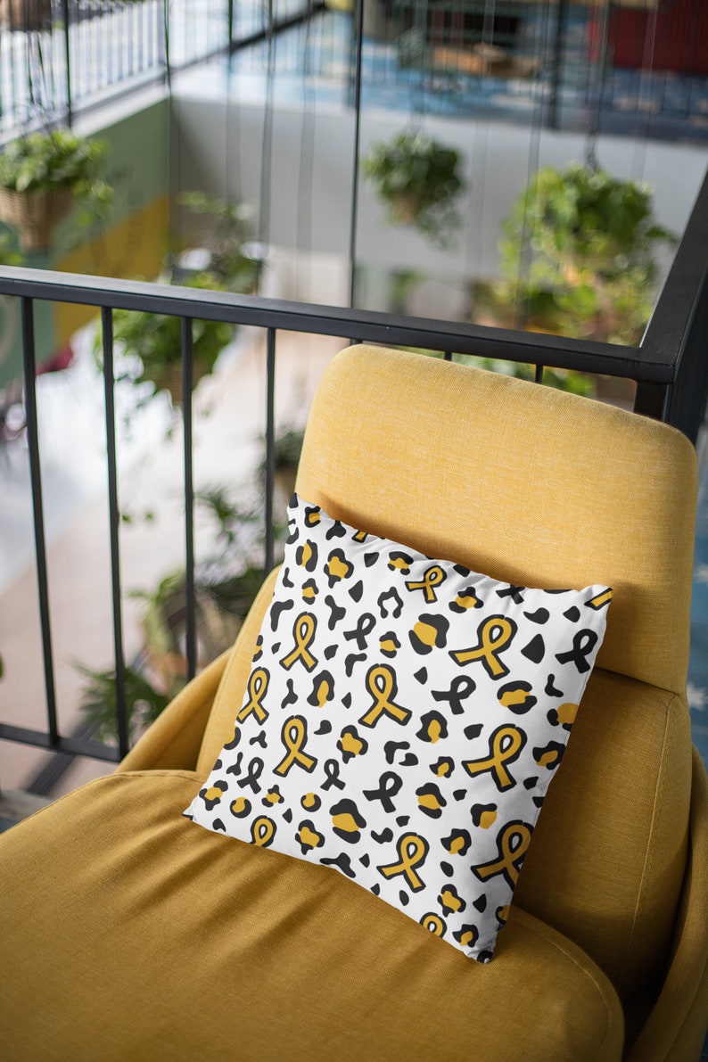 May include: A white throw pillow with a black and gold leopard print pattern. The pillow features a repeating design of yellow awareness ribbons.