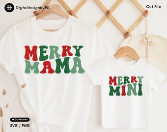 Op de afbeelding: Twee witte T-shirts met een kerstthema. Het grotere shirt zegt "Merry Mama" in rode, groene en witte blokletters. Het kleinere shirt zegt "Merry Mini" in dezelfde stijl.