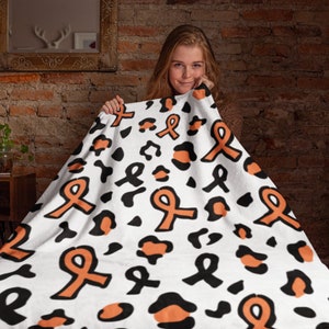 May include: A white fleece blanket with a black and orange leopard print and orange ribbon pattern. The blanket is being held up by a person.