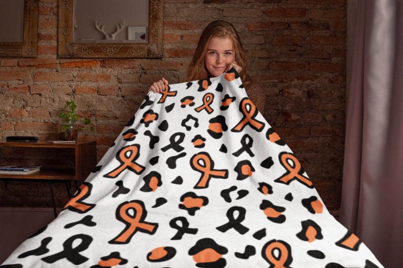 May include: A white fleece blanket with a black and orange leopard print and orange ribbon pattern. The blanket is being held up by a person.
