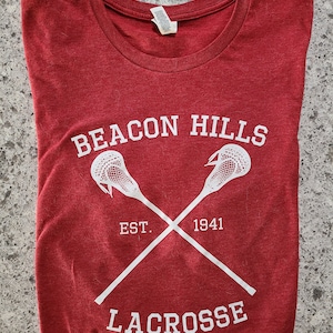May include: Red t-shirt with white text and a lacrosse stick graphic. The text reads "Beacon Hills Est. 1941 Lacrosse".