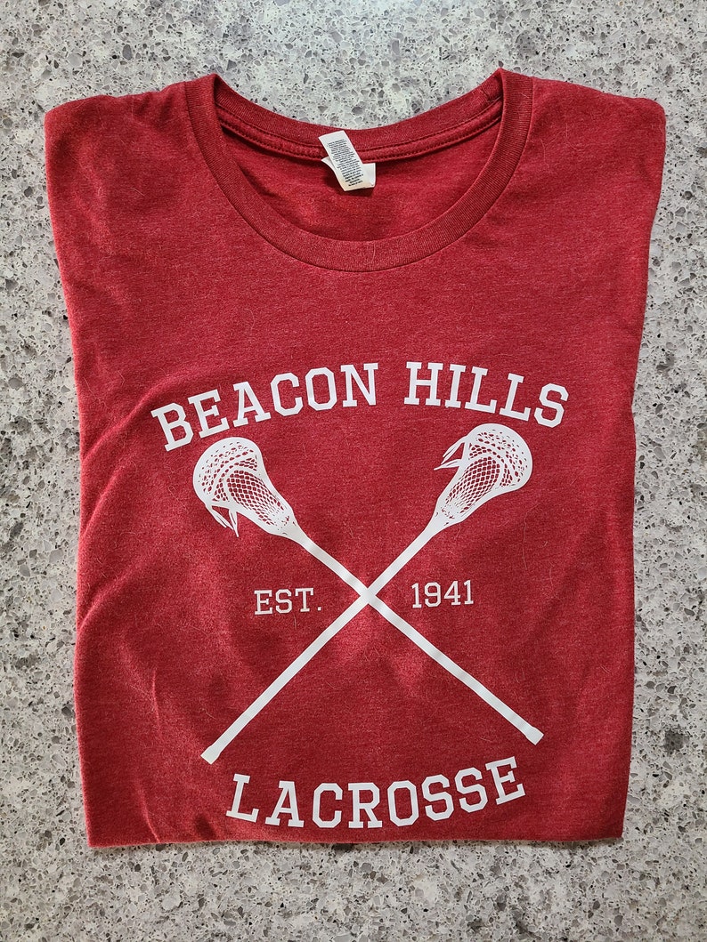 May include: Red t-shirt with white text and a lacrosse stick graphic. The text reads "Beacon Hills Est. 1941 Lacrosse".