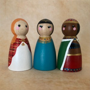 May include: Three wooden peg dolls, each with a different painted face and outfit. The doll on the left has a red and orange plaid dress and a white skirt. The middle doll has a blue dress with gold trim. The doll on the right has a green and red dress with gold trim.