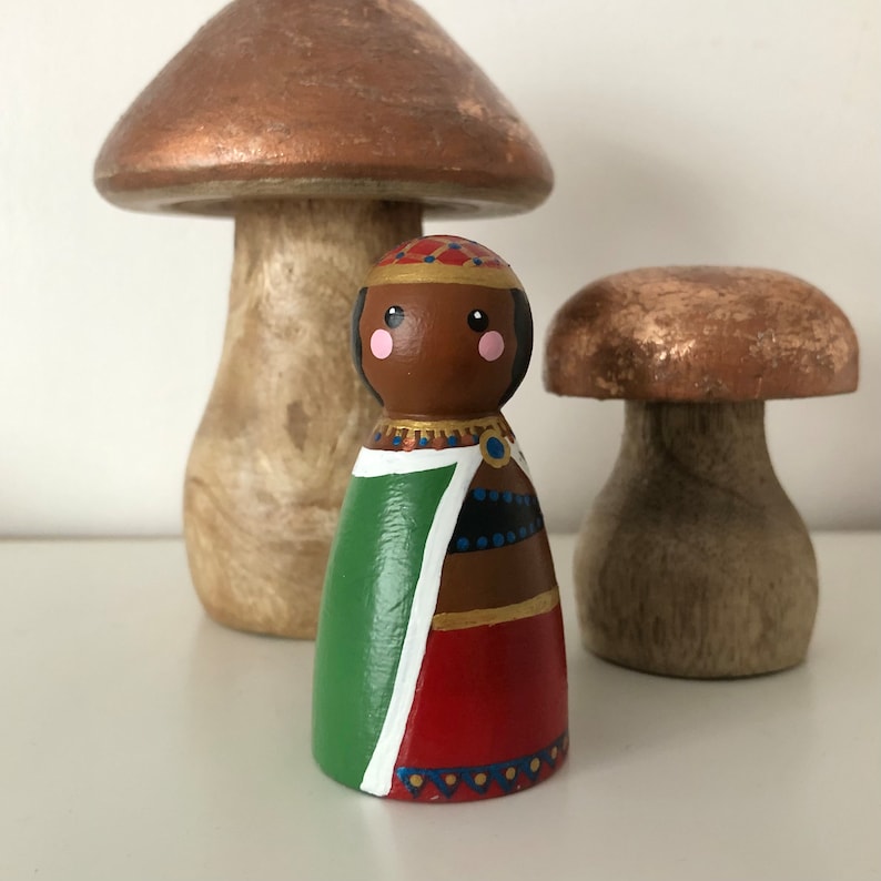 May include: A wooden doll with a brown face, green and red clothing, and a gold crown. The doll is standing in front of two wooden mushrooms.