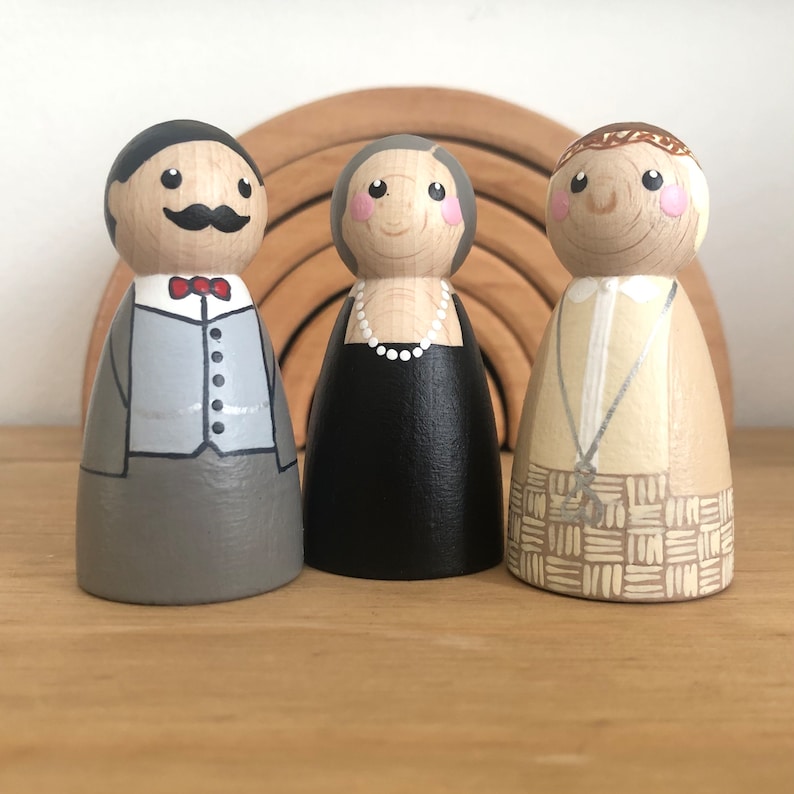 May include: Three wooden peg dolls, hand-painted with details. The doll on the left is wearing a gray suit with a red bow tie and a mustache. The doll in the middle is wearing a black dress with a white pearl necklace. The doll on the right is wearing a beige shirt with a silver necklace and a patterned skirt.