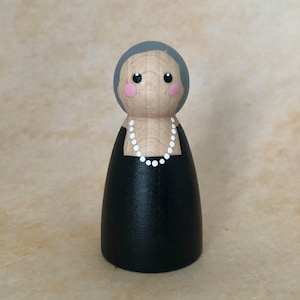 May include: A wooden peg doll with gray hair, pink cheeks, and black eyes. The doll is wearing a black dress with a white pearl necklace.