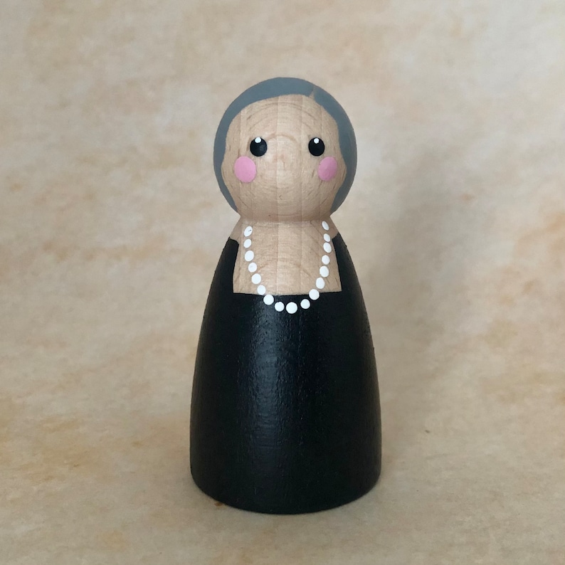 May include: A wooden peg doll with gray hair, pink cheeks, and black eyes. The doll is wearing a black dress with a white pearl necklace.