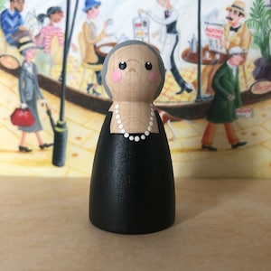 May include: A wooden peg doll with a gray hair, pink cheeks, and a black dress. The doll is wearing a white pearl necklace. The doll is standing in front of a painting of people in a cafe.