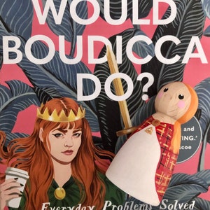 May include: A book cover with a pink background and a green leafy pattern. The title is "What Would Boudicca Do?" and the subtitle is "Everyday Problems Solved by History's Most Remarkable Women". A woman with red hair and a crown is holding a coffee cup. A wooden doll with a red and gold plaid dress is in the background.