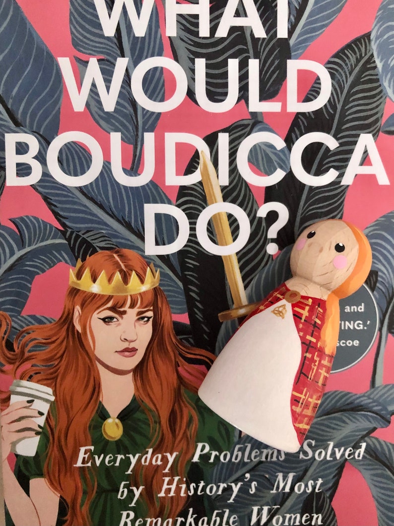 May include: A book cover with a pink background and a green leafy pattern. The title is "What Would Boudicca Do?" and the subtitle is "Everyday Problems Solved by History's Most Remarkable Women". A woman with red hair and a crown is holding a coffee cup. A wooden doll with a red and gold plaid dress is in the background.