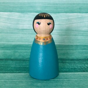 May include: A wooden peg doll with black hair, a gold crown, and a teal blue dress. The doll has a painted face with pink cheeks and black eyes.