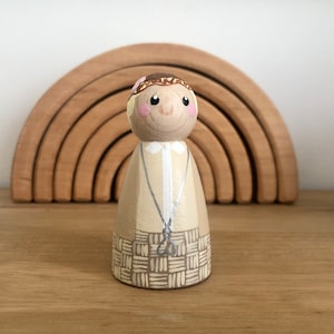 May include: A hand-painted wooden doll with a brown and beige outfit and a flower crown. The doll has a smiling face with brown eyes and pink cheeks.