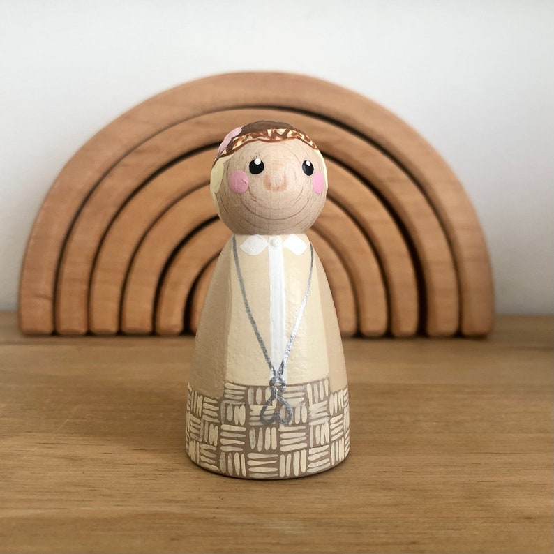 May include: A hand-painted wooden doll with a brown and beige outfit and a flower crown. The doll has a smiling face with brown eyes and pink cheeks.
