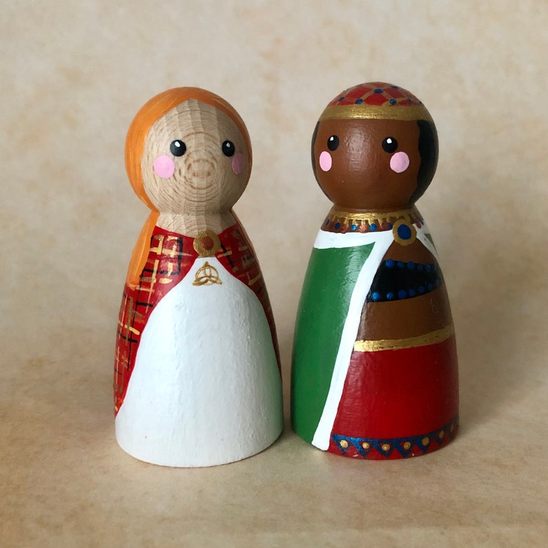 May include: Two wooden peg dolls, one with red and orange plaid and a white dress, the other with a green, red, and gold patterned robe.