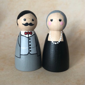 May include: A pair of hand-painted wooden peg dolls dressed in formal attire. The male doll is wearing a gray suit with a red bow tie and a black mustache. The female doll is wearing a black dress with a white pearl necklace.