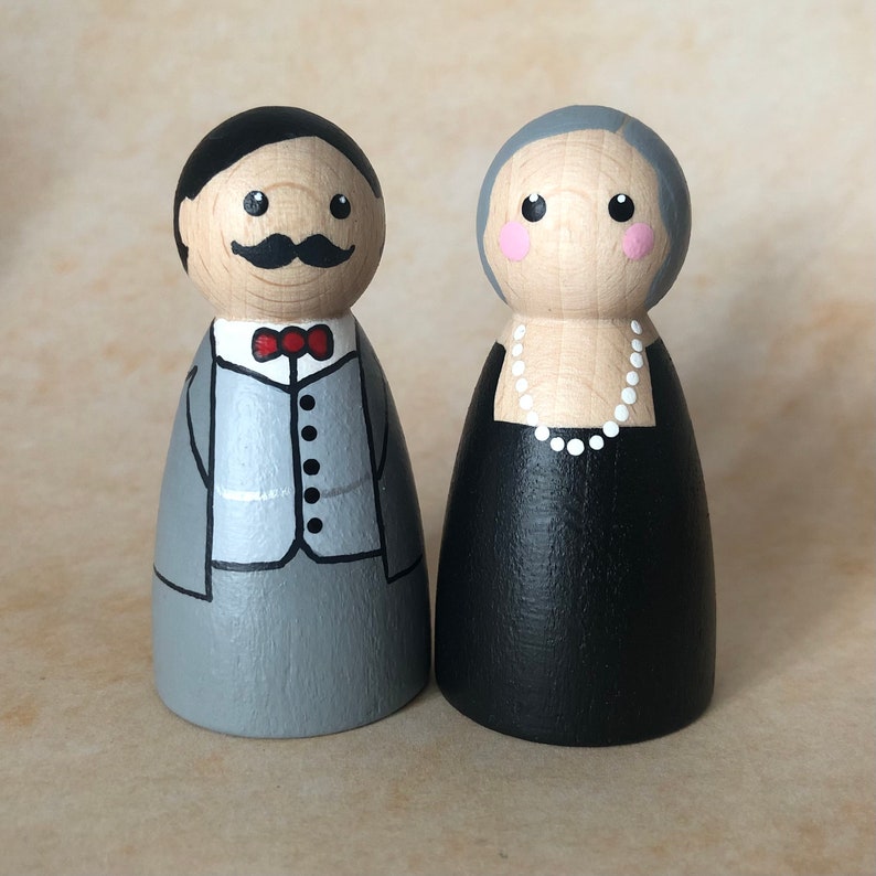 May include: A pair of hand-painted wooden peg dolls dressed in formal attire. The male doll is wearing a gray suit with a red bow tie and a black mustache. The female doll is wearing a black dress with a white pearl necklace.