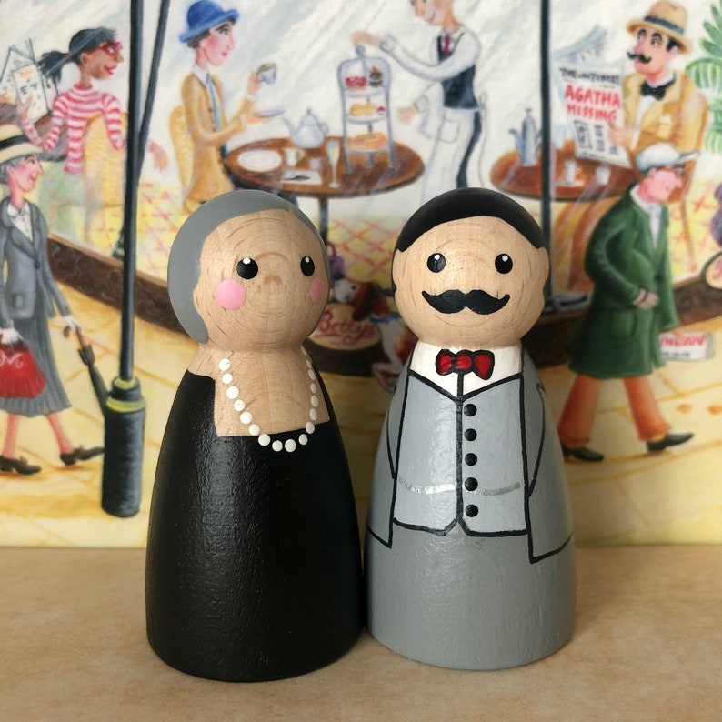 May include: A pair of hand-painted wooden peg dolls dressed in formal attire. The woman is wearing a black dress with a white pearl necklace. The man is wearing a gray suit with a red bow tie.