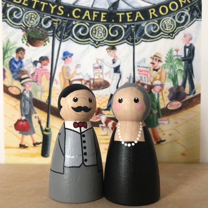 May include: A pair of hand-painted wooden peg dolls dressed in vintage clothing. The male doll is wearing a gray suit with a red bow tie and a mustache. The female doll is wearing a black dress with a white pearl necklace. The dolls are standing in front of a painting of a cafe.