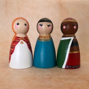 May include: Three wooden peg dolls, each with a different skin tone and wearing a different outfit. The doll on the left is wearing a red and white plaid dress with a white cape. The doll in the middle is wearing a blue dress with gold trim. The doll on the right is wearing a green and red dress with gold trim.