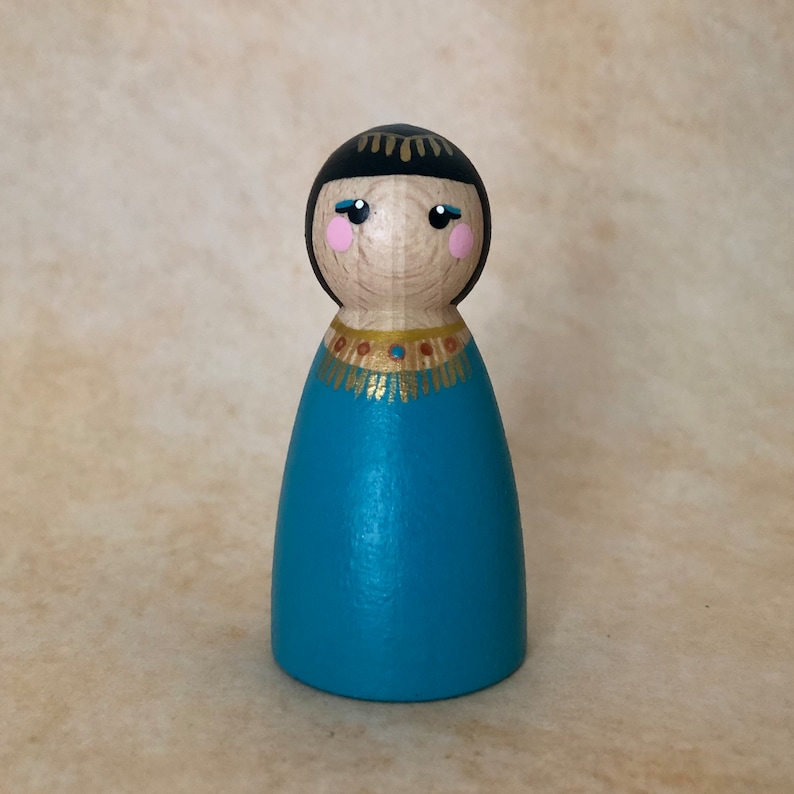 May include: A wooden peg doll with black hair, blue eyes, and pink cheeks. The doll is wearing a teal blue dress with a gold and brown beaded necklace.